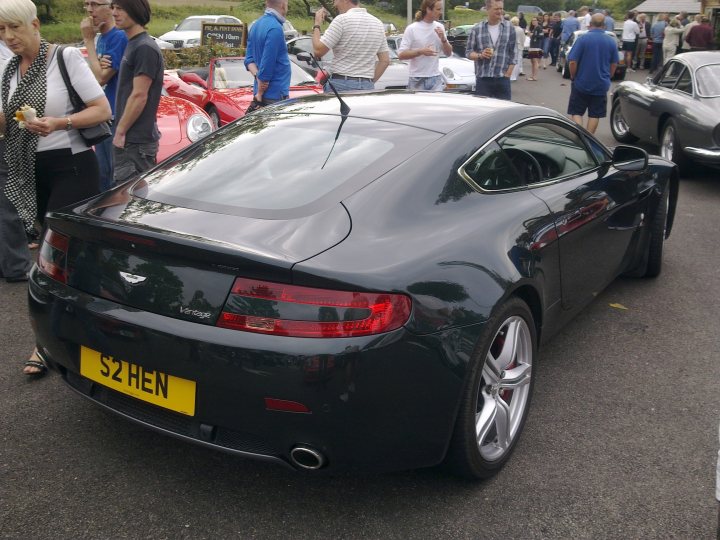 Pistonheads - The image shows a bustling scene where a black sports car with the license plate "S2 HEN" is parked in a parking lot filled with various vehicles and numerous pedestrians. The sports car is positioned to take up most of the frame, showing its sleek design and one of the rear wheels, while the others are in the background. The pedestrians, who appear to be at a public event, are scattered throughout the image, with some in the foreground and others in the distance. There is a mix of vehicles, ranging from other sports cars to trucks, suggesting a diverse crowd. The environment suggests an open, outdoor event or gathering.