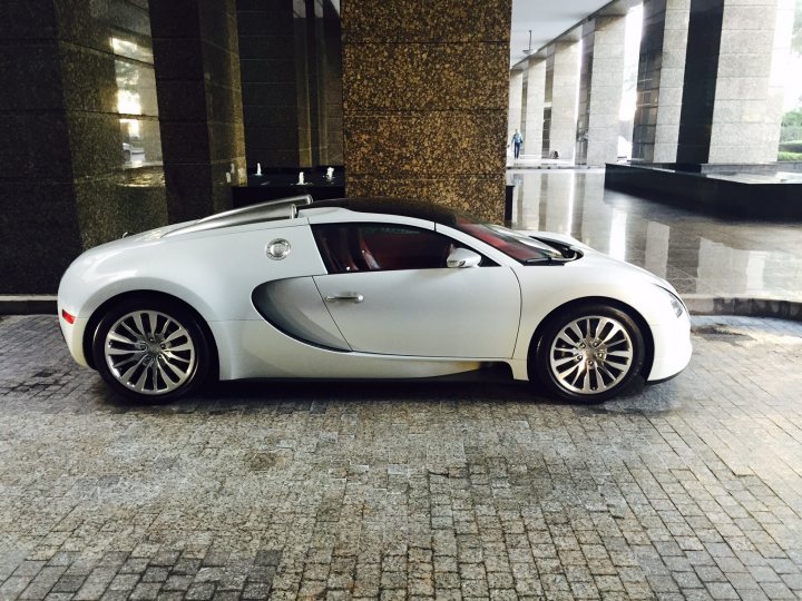 Middle East spotted thread - Page 77 - Middle East - PistonHeads - The image shows a silver Bugatti car parked at a curb. The car appears to be the Bugatti Veyron, characterized by its distinctive elongated shape featuring two cabin doors that also serve as air outlets, the quad tailpipes, the massive rear wing, and the alloy wheels. The Bugatti logo is prominently visible on the side. The car is positioned next to a street lamp, and there is a glimpse of architectural structures in the background, including what looks like a stairwell of a building. The sun seems to be shinning on the car, creating a bright reflection on the ground.