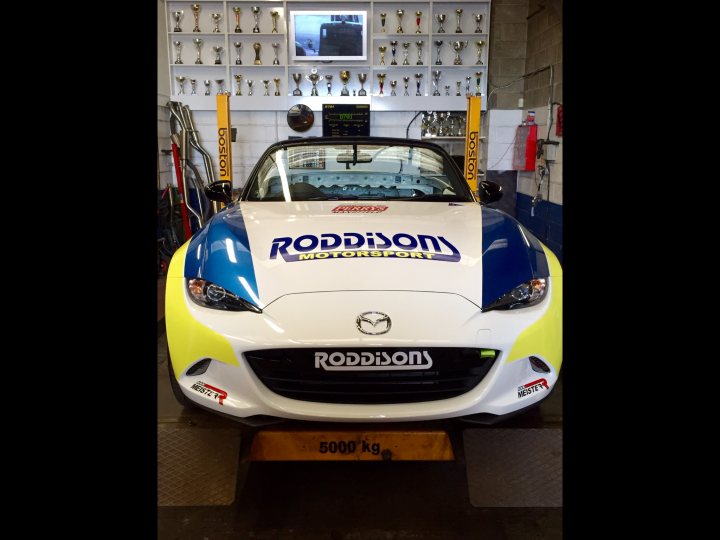 ND race car anyone? - Page 1 - Mazda MX5/Eunos/Miata - PistonHeads - The image shows a side view of a race car that is predominantly white with a blue stripe. The car displays the name "RLR RODDING MOTORSPORT" on the hood. A blue and yellow Rodadon license plate is visible in the foreground. The car is parked indoors next to emblems and other items that suggest victory or attainment, possibly indicating the car's racing history or awards. The setting includes various racing and automotive-related items in the background on a white tiled wall.