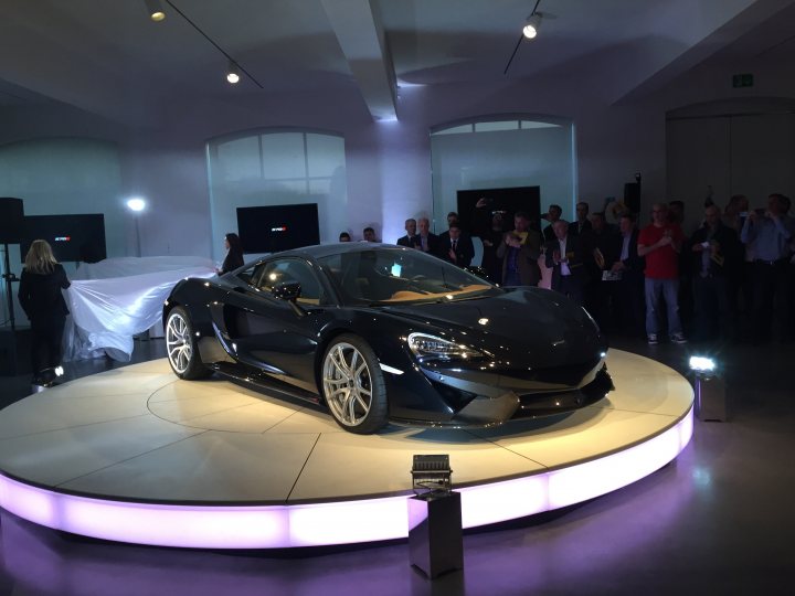 A car is parked in front of a building - Pistonheads - The image showcases a sleek, all-black electric sports car positioned in the center on a circular purple pedestal in an indoor showroom. This futuristic vehicle is designed with an aerodynamic body, evident from its streamlined silhouette and large wheel arches. The showroom itself is filled with artificial lighting and has a tiered layout featuring people on an upper balcony overlooking the main floor. The background is somewhat blurred, but it suggests a spacious room with high ceilings, further emphasizing the exclusivity and sophistication of the car.