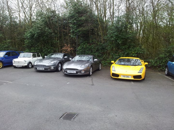 So what have you done with your Aston today? - Page 189 - Aston Martin - PistonHeads - This image captures a scene in a parking lot at what appears to be dawn or dusk. The lot is filled with cars, primarily older models. Two silver cars are parked close together, while a yellow sports car stands out by being separately parked. The vehicles display various colors and models, including two distinct hues of blue. The setting is surrounded by a lush tree line and a wide, grassy area. The lighting suggests a cooler part of the day.