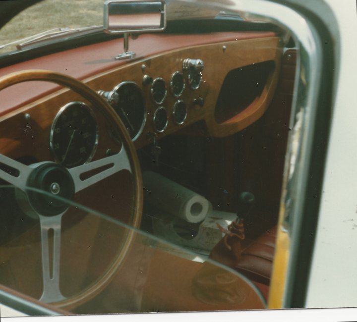 Early TVR Pictures - Page 33 - Classics - PistonHeads - The image shows a vintage car interior from the perspective of a passenger looking at a steering wheel. The steering wheel and dashboard have various instruments and gauges with many buttons and dials, typical of older cars. The upholstery appears to be a combination of wooden trim and leather seats, with a center console separating the driver and passenger seats. The photo itself is color, but aged, giving it a faded, nostalgic quality.