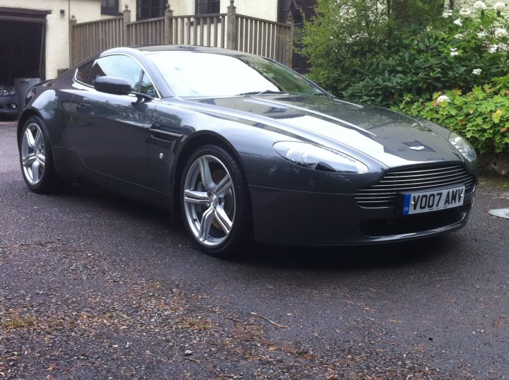 The Proper Colour for an Aston thread - silver, greys, black - Page 2 - Aston Martin - PistonHeads - The image features a convertible car, prominently bearing the front license plate Y007 AMY. The vehicle is parked on a driveway covered with gravel and tar, suggesting a residential setting. The car is the main subject, with the license plate clearly visible, adding a personal touch to the scene. There are green foliage in the background, providing a natural backdrop to the parked vehicle. The car appears to be dark in color, with the top up, indicating that it is not currently in use. The overall atmosphere of the image is serene and domestic.