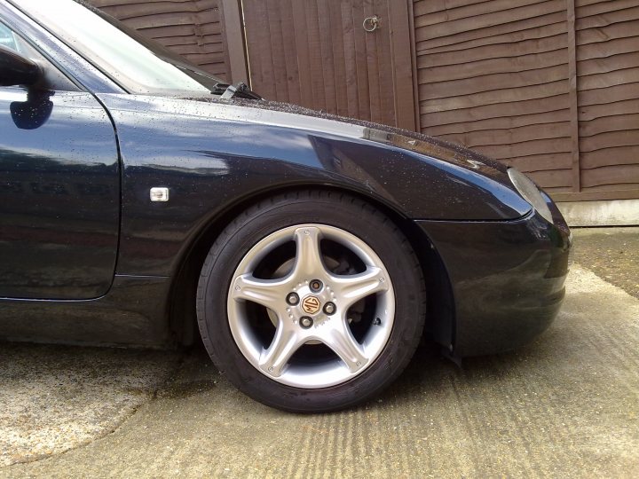 Mgf Pistonheads Cheap - The image showcases the left front wheel of a blue sports car parked on a concrete surface. The car features a prominent silver hubcap with the Audi emblem. On the inside of the wheel arch, mud splatters are visible, suggesting that the car has been driven through wet conditions. In the background, a wooden fence can be seen, indicating a residential or parking area. Overall, the photo gives a sense of the car's condition and environment.