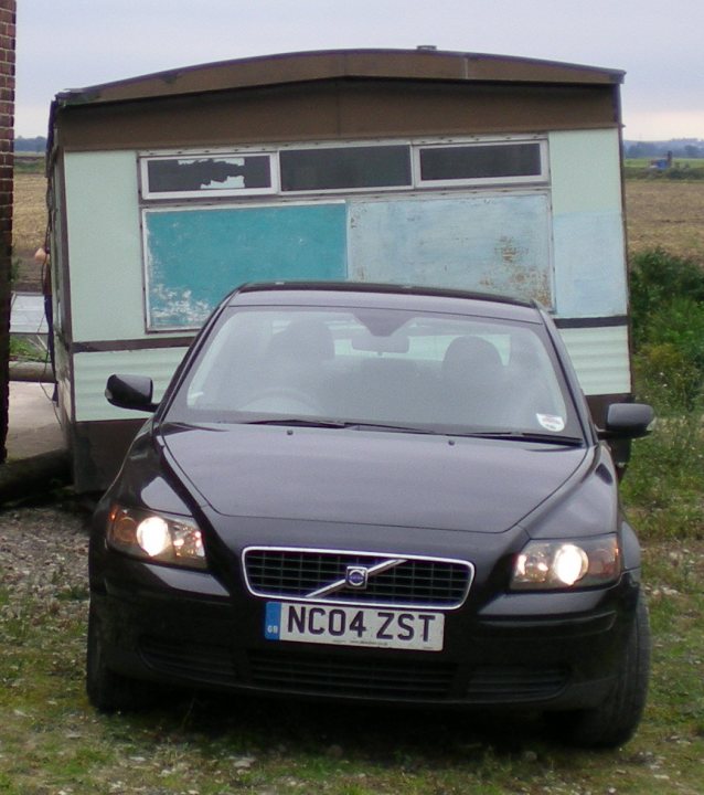 Volvo towing a caravan, erm...  ...your thoughts?? - Page 1 - Volvo - PistonHeads - This image features a dark-colored car parked in a grassy area to the left of an old, bright green and brown camper trailer. The car appears to be a modern model, but there's a sign on it which reads "NCO4 4ZST." The camper trailer has a slightly broken and discolored rear window, possibly from UV exposure, indicating it might be from an earlier era or not well-maintained. The sky is overcast, suggesting the photo may have been taken on a cloudy day.