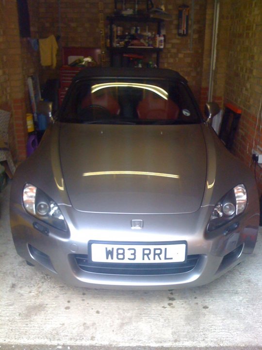 Pistonheads - This image features a sleek, silver sports car positioned inside an indoor parking space. The car, characterized by its distinctive front and side grilles and alloy wheels, faces towards the camera. The vehicle's license plate reads "W83 RRL". Behind the car, a brick wall is reflected, suggesting a mirror or similar surface. The space also contains various objects such as a shelf, a refrigerator, and some chairs, indicating that the structure might be used for multiple purposes other than parking.