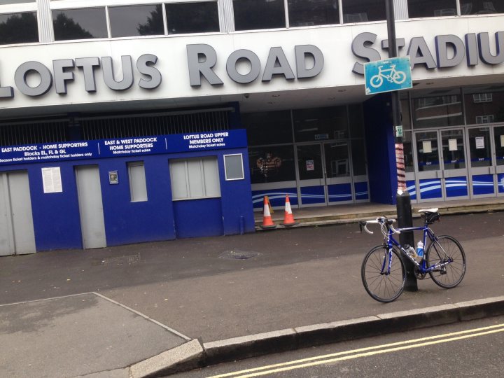 The "Photos From Today's Ride" thread... - Page 150 - Pedal Powered - PistonHeads - The image displays the exterior of a building with a blue and white striped facade. The signage on the building reads "LOTTUS ROAD STADIUM" in large, bold black letters. A blue and white bicycle is parked to the right of the image, and there are a few icons or signs affixed to the building near the bicycle, one of which shows a bicycle and the text "CICLE PARKING." In front of the building, there is a paved area leading to a set of doors and a small triangular figure on the ground. There's also a tree visible near the bicycle. The style of the image is a candid photograph taken during the day.