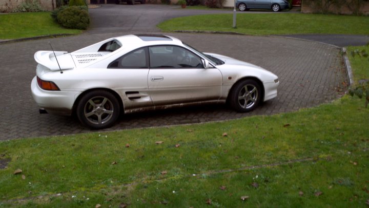 MR2 specialist near Sherborne? - Page 1 - South West - PistonHeads - In the image, there is a white sports car parked on a paved driveway adjacent to well-maintained green grass. The car is a two-door coupe with a rear-header spoiler which adds to its racing-inspired look. The vehicle is parked, ready to drive, with its side facing the camera. In the wider frame of the image, there is another smaller car, possibly a compact, visible in the background. The scene suggests a suburban residential setting.