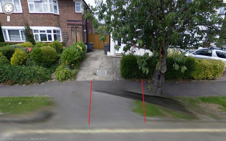 Widening the dropped kerb (Driveway) - Page 1 - Homes, Gardens and DIY - PistonHeads - The image is a fisheye, panoramic photograph capturing a street scene. The banding effect of the lens is evident, giving the photograph a distorted, panoramic quality. The street in the foreground appears asphalt with distinctive red and yellow lines, possibly indicating parking or no parking zones. On the right side of the photo, there's a glimpse of residential architecture, including a red brick building with a brown door, a brick wall, and trimmed, green hedges lining the path. The tree ranks prominent in the background, providing a natural contrast to the urban setting.