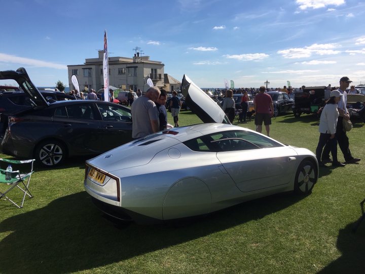 RE: Volkswagen XL1: Spotted - Page 3 - General Gassing - PistonHeads - This image captures a lively scene at a car show. A futuristic, silver electric car is the focal point, parked beautifully on the lush green grass. The car's sleek design and the word "electric" on its side suggest it's an innovative vehicle. Surrounding the car, a group of people have gathered, examining the car and discussing its features. They are standing around a few more cars that are parked nearby, adding to the atmosphere of the event. The backdrop is a beautiful, sunny day that has further brightened the scene.