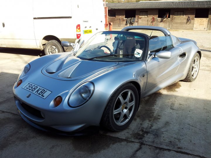 S1 Original Silver(s) - Page 1 - Elise/Exige/Europa/340R - PistonHeads - The image shows a sleek convertible sports car parked in a covered area or driveway next to another vehicle. The car is predominantly silver in color, with a visible license plate and a partial view of its front left wheel. It appears to be parked on a paved surface. In the background, you can see a white van and stables or wooden structures that suggest a rural setting. There are no visible texts in the image.