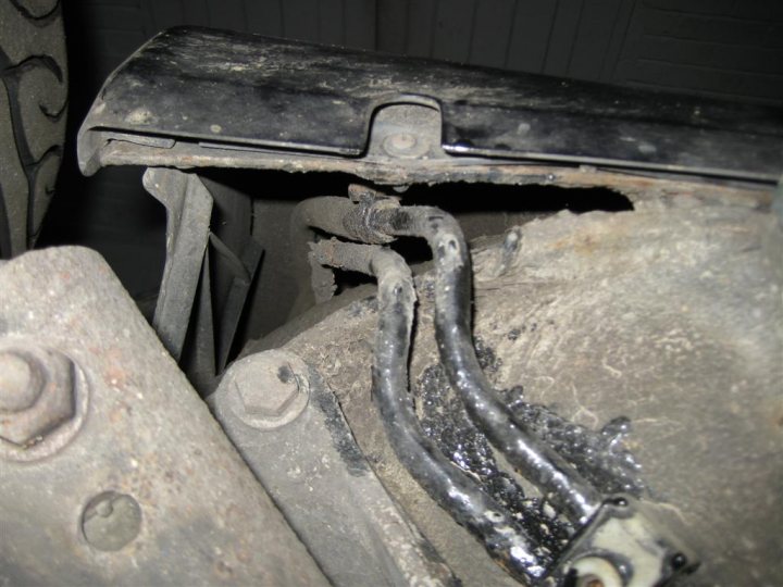 A close up of a machine on a train track - The image shows a close-up view of the underside of a truck, specifically focusing on its suspension system. There is a noticeable absence of a bumper or fender, revealing the components underneath such as springs and struts. The area around the shocks has visible rust and corrosion, indicating wear and tear or potential damage. A few drops of grease can be seen around the exposed metal components.