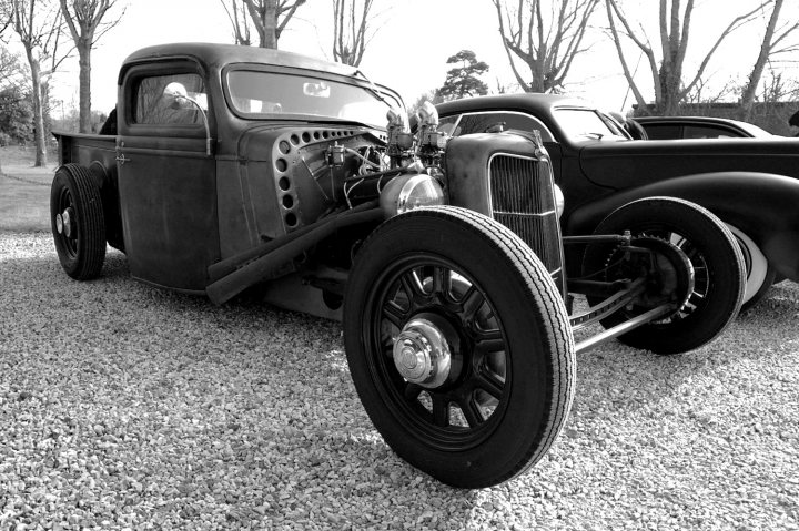 Goodwood Breakfast Club - Sushi Sunday - photos - Page 1 - Goodwood Events - PistonHeads - The image is a striking black and white photograph capturing the classic elegance of a vintage vehicle. The main focus is an antique pickup truck, which exudes an aura of timeless charm. The truck is parked amidst a gravel-strewn landscape, with trees standing tall in the background, adding to the rustic setting. Its robust and sturdy body is accentuated by its large, round tires and a small louvered hood, which are characteristic of such vehicles. The texture and contrast of the photo, being black and white, bring out the details of the truck and the environment, making it a captivating piece of art.