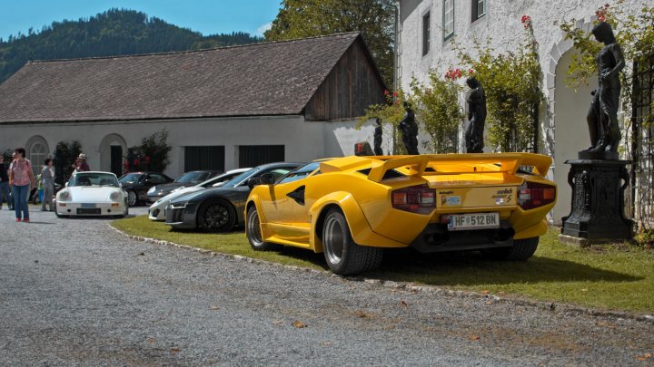 Null's ultimate Dream Car Thread - A CV8 Monaro in Austria - Page 9 - HSV & Monaro - PistonHeads UK - The image depicts an outdoor setting where various luxury cars are parked. There's a yellow sports car in the foreground, and several other high-end cars, including a Lamborghini, are visible in the background. The cars are on gravel, suggesting they might be at a private event or a racing circuit. There are people present, possibly attendees of an event or spectators. A building with a wooden facade and a roof garden is in the background, providing a contrast to the hard, metallic surfaces of the vehicles. The sky is clear and blue, indicating fair weather.