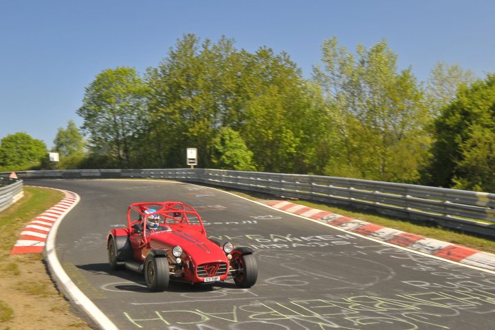 Work Pistonheads Part - The image presents an exhilarating scene on a race track. At the center of the frame, a red sports car is making a turn, leaning low on its suspension as it is skillfully maneuvered by a driver. The track is bordered by a grassy area, indicating the boundary of its closed circuit. Surrounding the track, trees provide a natural backdrop to this intense racing activity.