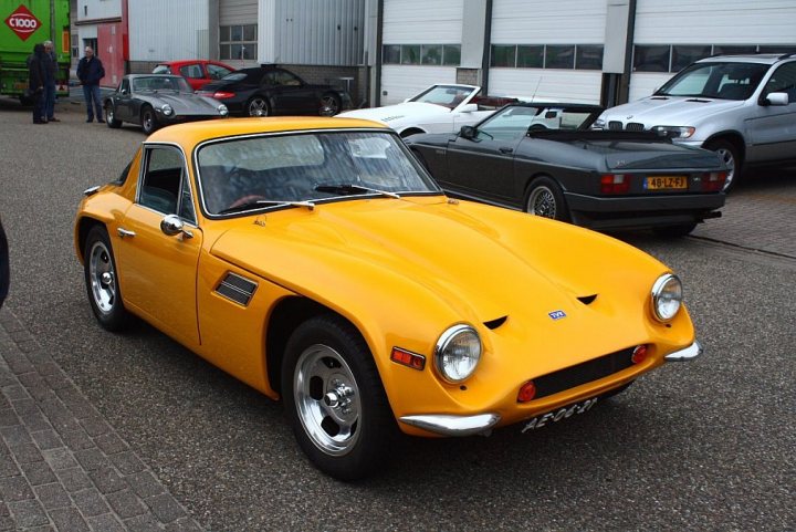 Early TVR Pictures - Page 81 - Classics - PistonHeads - The image presents a row of vintage sports cars parked on an asphalt surface, set against a backdrop of a building with a garage door. Dominating the scene is a bright yellow sports car with distinct wedge-shaped front and rear spoilers, indicating a classic style often associated with the 1970s. The car's metallic hue stands out amidst the more subdued colors of the surrounding vehicles. The others cars, parked haphazardly, include models with designs that suggest they come from different eras but share similar aesthetics. The scene conveys a sense of nostalgia and appreciation for classic automotive design.