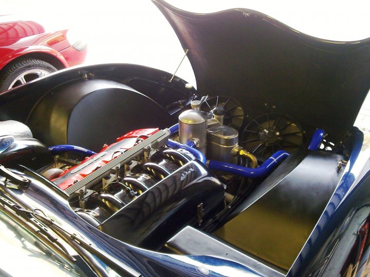 Pistonheads - The image displays the open hood of a sports car, revealing a complex engine with various components visible. The engine is large, featuring multiple cylinders and a header that appears to be integrated into the bodywork of the vehicle, which is a striking metallic blue with black accents. Nearby, a red car glimmers under a steep angle, adding contrast to the scene. The view is from a slightly elevated perspective, capturing the mechanical beauty of the engine while also showcasing the design of the car.