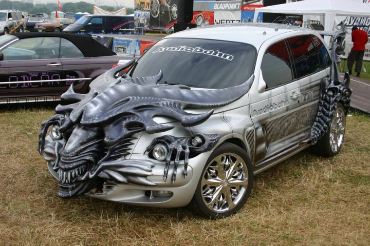 Pistonheads - The image features a silver-colored BMW SUV parked on a grassy area amidst other cars. The SUV stands out with its gray dragon-like hood decoration. This dragon-like car sculpture is also known as a "chopper car design," often seen at car shows for customized vehicles. The presence of the dragon hood adornment suggests that it might be a part of a car customization or a prop for an event, such as Halloween or a themed party. The spiky, fantastical dragon head adds an element of surprise and creativity, making the car unique and eye-catching in comparison to standard vehicles. In the background, there's a bustling auction area with various other cars and people, indicating that the setting might be a car show or an auction event.