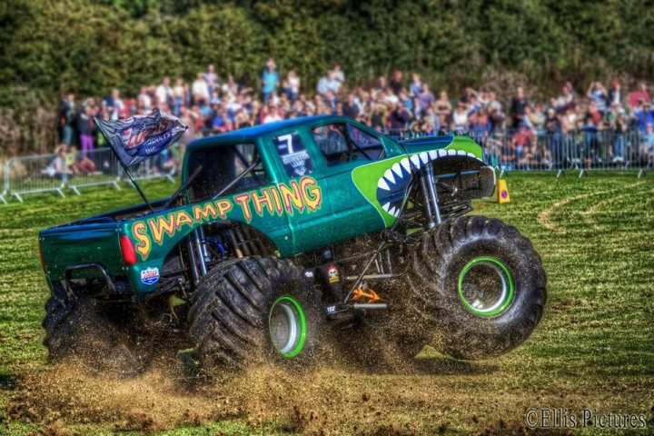 A green and black truck is parked in a field - Pistonheads - In the image, a monster truck wheeled out a dump of earth, indicating it has flipped. The truck is predominantly green and features a giant shark or whale fin on the back, with a banner in a large font that reads "SWAMP THING". The setting appears to be a typical monster truck event, with a crowd of spectators visible in the expanse behind the truck.