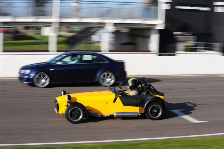 Not enough pictures on this forum - Page 1 - Caterham - PistonHeads - The image presents an engaging scene on a race track. At the forefront, a yellow three-wheeled car is seen making a sharp turn around the race track. This car appears to be smaller in size as compared to the car following it. 

Behind the three-wheeler, a larger, dark-colored car is visible, driving at a steady pace. The contrast in the vehicle colors against the track and overcast sky in the background. 

In the periphery, we can see a person wearing a helmet, possibly indicating an active participant or a crew member on the track. 

Overall, the image conveys a sense of speed and competition, with the smaller three-wheeler taking the lead over the larger car.