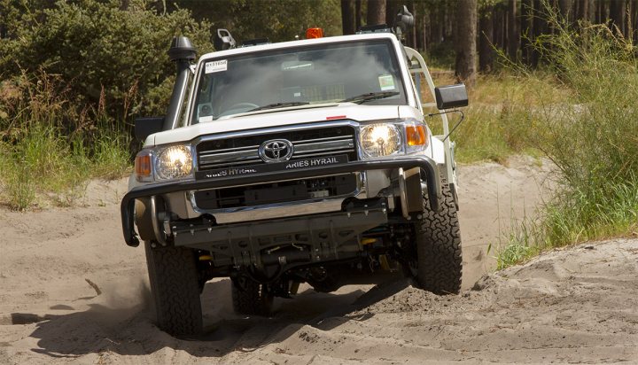 4WD Rental in Perth? - Page 1 - Australia - PistonHeads - This image captures a Toyota Tacoma Trail truck, which is well-suited for off-road adventures. The truck is driving through a dirt road that appears to be quite sandy or rocky, indicating that it's off the beaten path. The Tacoma is equipped with a snorkel and hoop bumpers, which are functional modifications for rough terrain. There are no visible texts or license plates, and the environment around the truck is a mixture of dirt and greenery, suggesting it might be in a camping or outdoor recreational area. The camera angle is from the driver's side, giving a sense of the truck's movement and the rough conditions it's navigating.