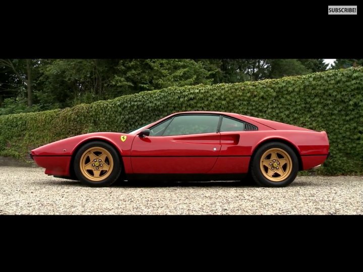 308/328 Values - Page 14 - Ferrari Classics - PistonHeads - The image displays a vibrant red Ferrari sports car parked on a gravel surface. The car is sleek and has a low-profile design with silver rims contrasting against the black of the tires. It's positioned with a side profile, showing the distinctive shape and timeless design of the Ferrari, which includes a large air intake at the rear behind the engine/spoiler area. The background features a dense hedge, indicating a private or park-like setting. There is a subscribe bar at the bottom of the image.