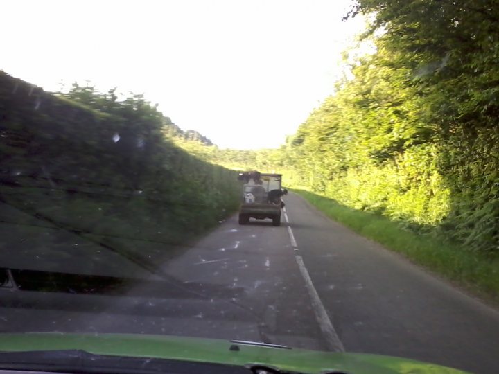 Best bits of roads around Exmoor - Page 1 - South West - PistonHeads - The image appears to be taken from the perspective of the driver's seat inside a vehicle. The car has a green hood, and there's an additional green object to the left side of the front windshield. The driver is visible, sitting behind the steering wheel. The windshield is slightly dirty with streaks across it. A large, leafy green bush or hedge borders the road to the left. There are also open fields on the right side of the road, leading to a rural, country setting. The sunlight is penetrating the scene, suggesting an outside temperature that is somewhat warm.