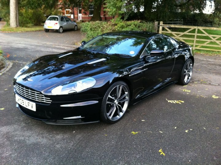 I'm grinning! - Page 1 - Aston Martin - PistonHeads - The image presents a sleek, black Aston Martin car parked in a driveway on a paved surface. The car is in front of a yellow gate with a wooden structure behind it. There are two other cars visible in the background, one of which is silver and the other is white, indicating a street or parking area nearby. The surrounding area includes elements of nature, such as leaves scattered on the pavement, adding a touch of the outdoors to the scene.