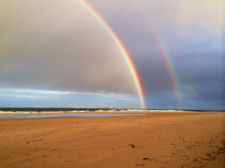 Just for fun random camera phone pictures you're happy with. - Page 11 - Photography & Video - PistonHeads - The image features a sandy beach with two rainbows arching over it. The rainbows, appearing in the middle of the image, are vivid and colorful. The sand below is a rich brown, and the beach extends from the middle to the bottom of the image. In the background, the ocean meets the sky, providing a serene backdrop to the vibrant rainbows. The weather appears to be cloudy, as gray clouds fill the upper portion of the sky.