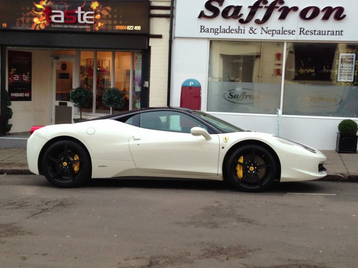 Spotted 2013 - Page 1 - Northamptonshire - PistonHeads - The image showcases a pristine white Ferrari parked on the street, positioned in the foreground with the cityscape and buildings in the background. The Ferrari is sleek and modern, with distinctive black tires and a yellow license plate attached to its rear. The street has a cobblestone texture and is lined with various shops and restaurants, adding to the urban atmosphere. The "SAFFRON" sign on the building suggests the presence of a restaurant named Saffron that serves both Bangladeshi and Nepalese cuisine. The clear sky and daylight illumination suggest that the photo was taken during the day.