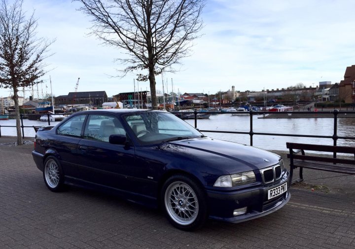 RE: BMW M3 (E36) | PH Used Buying Guide - Page 3 - General Gassing - PistonHeads - The image is a photograph of a car parked on the side of a street next to a body of water. The vehicle appears to be an older BMW model, characterized by its angular headlights and distinctive kidney grille. It's painted in a deep blue color with silver rims. There are no visible texts or license plates on the car. In the background, there is a clear view of calm waters where a few boats are docked, suggesting that this might be a harbor area. The sky is overcast, and the overall scene conveys a serene and possibly cool climate.