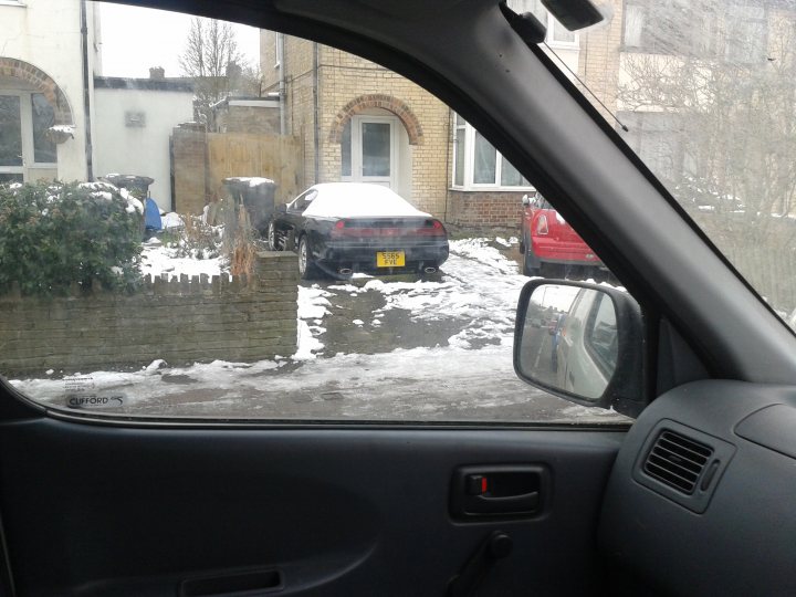 Classics left to die/rotting pics - Page 206 - Classic Cars and Yesterday's Heroes - PistonHeads - This is a rear-seat perspective from inside a car, showing a residential street scene. In the foreground, there is a black car and a red car parked next to each other. The focus of the image is through the car window, emphasizing the cold winter weather with snow-covered ground and brick walls. The mirror on the car's side reflects more of the street scene, providing an additional perspective.