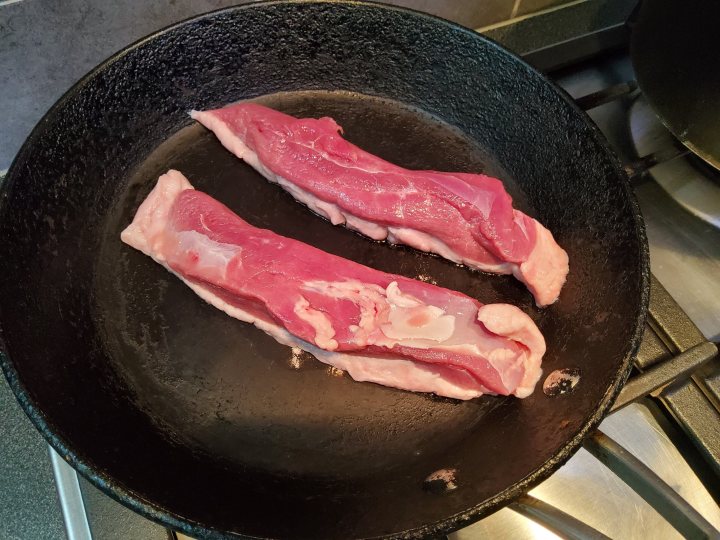 The image captures a close-up view of a sizzling pan on a stove, containing two large, uncooked pieces of meat. The meat appears to be steak or beef cutlets, and it's seasoned with salt, giving the surface a white, speckled appearance that contrasts with the darker hue of the meat itself. The pan is black and seems well-heated, suggesting that the cooking process has just begun or is paused momentarily. The stove beneath the pan is not fully visible but appears to be a conventional gas or electric stove. There's no text present in the image.