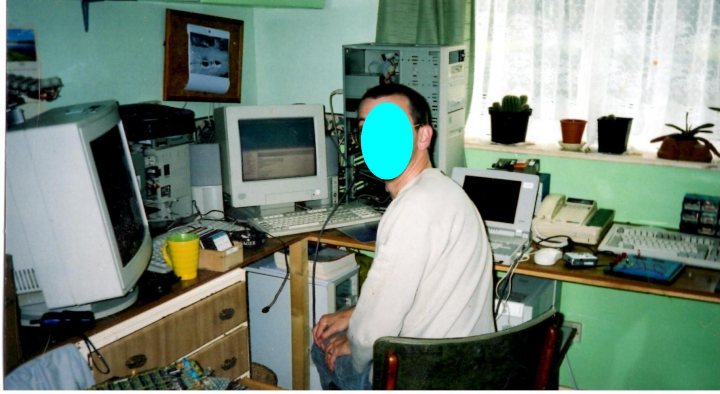 A man sitting in front of a laptop computer - Pistonheads - The image captures a homely workspace, dominated by a man seated at a desk. He is engrossed in work on a computer, his focus apparent even through the photo. The room is filled with various tech gadgets, with several computers and monitors scattered around him, adding to the virtual chaos of the workspace. A green chair adds a touch of color to the room, providing a stark contrast against the white walls. The room also houses a variety of plants, contributing to a soothing ambiance. The desk area is well-lit, with at least four sources of light visible in the photo. The man in the picture is of medium height, sporting glasses and a white long-sleeve shirt.