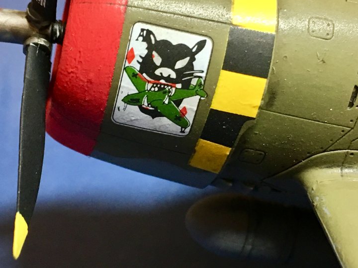 P-47 Thunderbolt “Razorback”, Academy 1:72 - Page 1 - Scale Models - PistonHeads - The image shows a close-up of a model airplane propeller, which has a graphic of a cartoonish green cat with the word "ACE" in the center. The cat is styled in a whimsical manner, with a black face, red eyes, and yellow fur accented with black spots. The model airplane appears to be a vintage fighter jet, painted in muted tones of brown and green, complemented with yellow stripes. The propeller sets against a blue background.