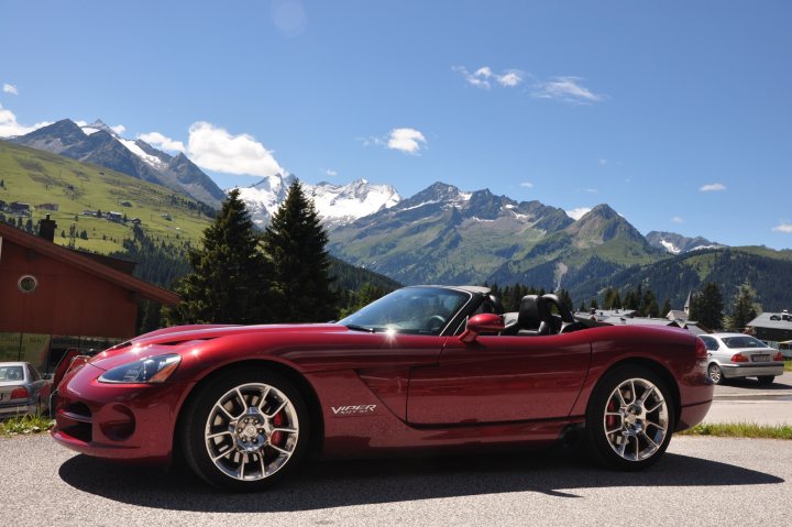 Hi Guys - Page 1 - Vipers - PistonHeads - The image captures a vibrant red sports car parked on a gravel road, its sleek design contrasting with the rugged landscape around it. The car, with its two occupants, is the focal point of the scene, positioned centrally in the frame. It faces towards the left, giving viewers a clear view of its polished wheels and emblematic design. Behind the car, a picturesque mountain range stretches across the horizon, their peaks adorned with patches of snow. On the ground, small trees dot the landscape, their stick thin forms adding a touch of texture to the otherwise smooth gravel. The scene exudes a sense of adventure and freedom, embodied by the open road and the cars readiness for the journey ahead.