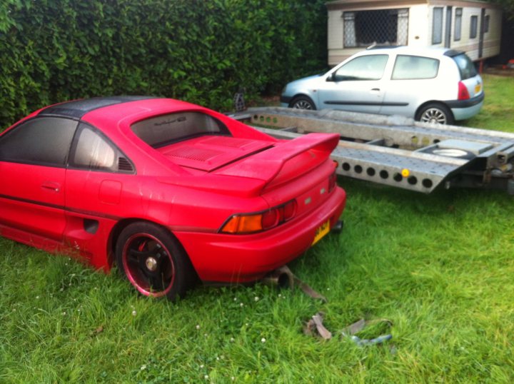 A red truck is parked in a field - Pistonheads - The image shows a scene of two wagon-style hatchback cars parked in the grass. A red sports car is prominently visible, parked as if abandoned with its hood up. Next to it is a grey-blue hatchback, which appears to be in better condition. A trailer is also present, partially obscured by the red car. The setting appears to be amidst a line of hedges, possibly serving as a boundary or a hedge maze. The overall impression is one of an unusual parking scenario, with a suggestion of abandonment or disrepair.