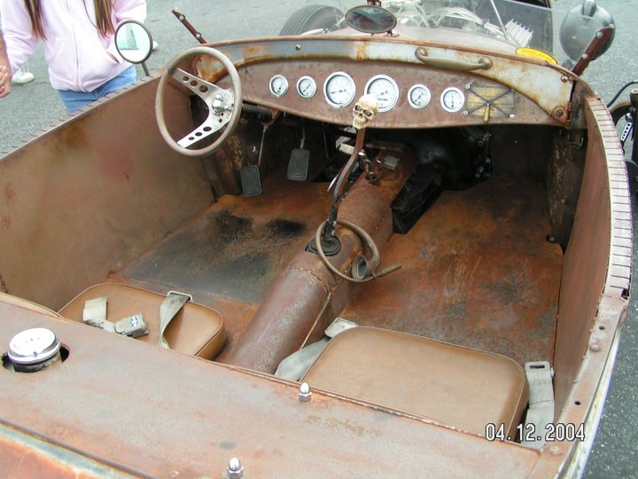 Pistonheads Rat Understand Doesnt - In the center of the image, there is an old, rusted car with an open hood, revealing its interior. The car is in a state of disrepair, with various parts missing or damaged. The driver's side is quite dirty, and there is a skull mounted on the dashboard of the car. In the background, there is a person partially visible, although it's unclear if they are interacting with the car or just passing by. The vehicle is predominantly brown and has a distinctive, rounded shape. The image does not contain any visible text.