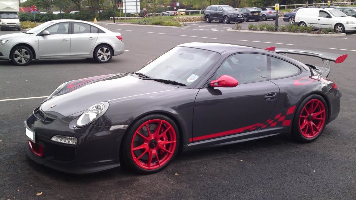 New Toy! - Page 1 - Porsche General - PistonHeads - The image shows a modern-looking sports car parked on the side of a road. The car's body is predominantly black, with the hood and part of the front fender accentuated in red and white. The vehicle features distinct red accents around the wheel arches and thick black stripes running down the side of the car. It has a dynamic design with pronounced curves and bulges, suggesting aerodynamic features for improved performance. There are additional details such as a sunroof and two red mirrors. The car has a large rear spoiler, indicating fast speeds and additional lift and downforce at the rear. It's stationed on asphalt, lined with white boundary markers, and the background reveals other parked cars and a clear sky.