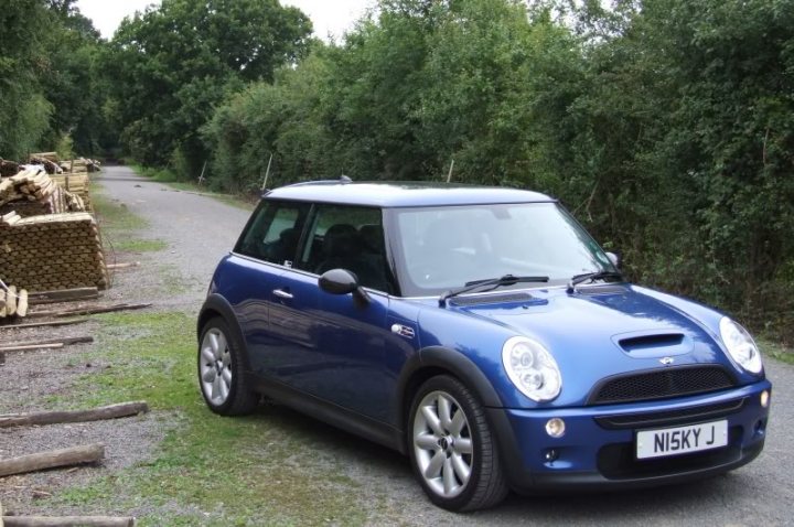 Pistonheads - The image depicts a small blue car parked on the side of a road surrounded by a lush green forest. The car is a Mini Cooper, identifiable by its distinctive design and badging. There are stacks of firewood nearby, suggesting a rural or suburban setting. The sky is partially cloudy, and there are trees with foliage in the background. The overall scene conveys a tranquil, outdoor location.