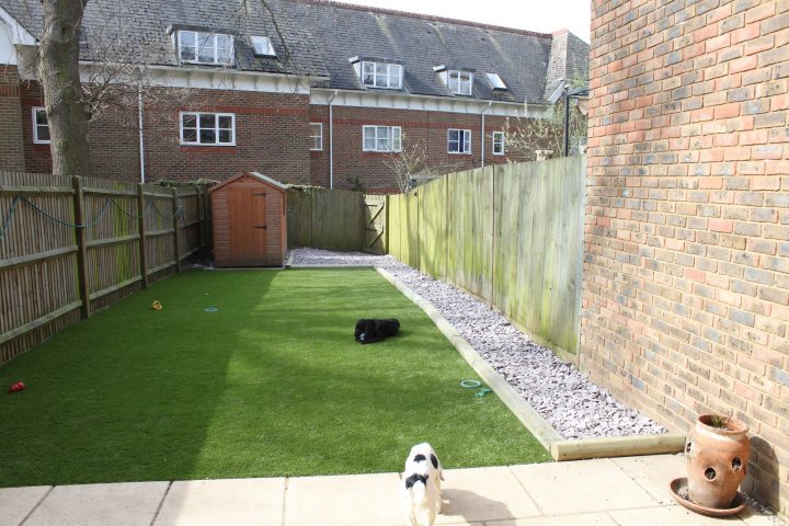 Ideas for a decent garden with dogs - Page 1 - All Creatures Great & Small - PistonHeads - The image depicts a serene outdoor spaces, dominated by the presence of a well-manicured backyard. Centered in the yard is a vibrant green artificial grass, a tranquil spot for a dog that rests comfortably on top of it. The dog, a medium-sized breed, lies on its side, seemingly relaxed, adding a sense of warmth and domestication to the scene. A red ball, possibly a toy, is visible on the grass, adding a splash of color to the green expanse.

On the left side of the yard, there's a wooden shed and a fence running along the yard, providing a boundary between different segments of the landscape. The shed's color contrasts with the green of the grass, while the fence is made of wood, complementing the overall outdoor setting.

The backyard is enclosed by a brick wall, partially obscured by a tree on the left. The brick wall, with its sturdy and timeless appearance, adds an element of permanence and structural stability to the scene. The tree, providing shade to the yard, enhances the natural aspect of the space.

Overall, the image portrays a calm and peaceful