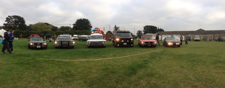 A bunch of trucks are parked in a field - Pistonheads - The image depicts an outdoor scene where various emergency vehicles are parked in a row on a grassy field. From left to right, there is a fire truck followed by two pickup trucks, and then two more vehicles that are not clearly identifiable from this angle. There is a group of people standing in front of the vehicles, and in the left foreground, another person can be seen walking away from the vehicles. The sky is overcast, suggesting a cool, possibly cloudy day. The setting appears to be a public space, possibly during an event or demonstration, as indicated by the size of the crowd and the abundance of equipment.