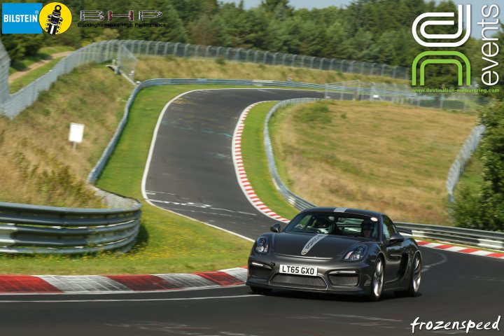 Porsche Cayman GT4  - Page 5 - Readers' Cars - PistonHeads - The image is an action-packed photograph of a single-occupant sports car in motion on a winding road. The car has visible license plates and is positioned with one wheel on a red curb, suggesting rapid acceleration. The environment shows a road that curves sharply to the left and then right, with a fence and grass boundary on the right. The surrounding landscape features dense green vegetation and a blue sky. There are logos in the top left corner and another on the car itself, indicating possible corporate sponsorship or association with events or organizations. The image is vibrant and conveys a sense of speed and racing through its dynamic composition and the car's motion blur.