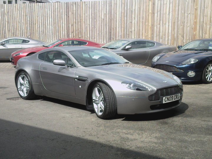 Just got my 09  vantage - Page 1 - Aston Martin - PistonHeads - The image shows a parking area with a line of sports cars parked side by side. Some cars are red, others are gray, suggesting a variety of models or manufacturers. The ground appears wet, and the area is slightly sloping. The cars are positioned at an angle to the camera, showcasing their design. There seems to be a fence or barrier in the background behind the cars.