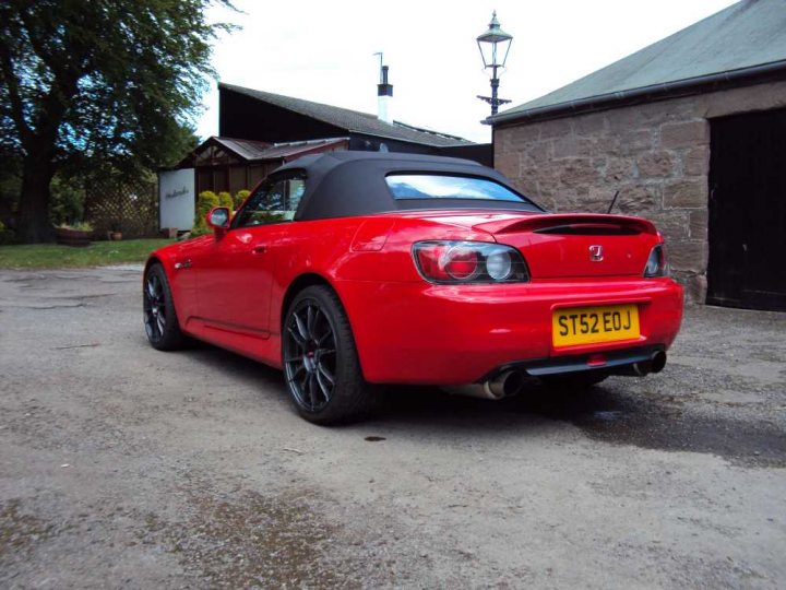 Pistonheads - The image shows a red sports car parked on a patchy dirt ground. The car is a two-door convertible with the top down, revealing the driver's cockpit. It features a drop-top design, black door handles, gray rims, and a yellow license plate that reads "ST52 EOJ." In the background, there is a large, stone building, a smaller building, and a metal light pole. The light and shadows indicate that the photograph was taken during the day. There are no visible people in the scene.