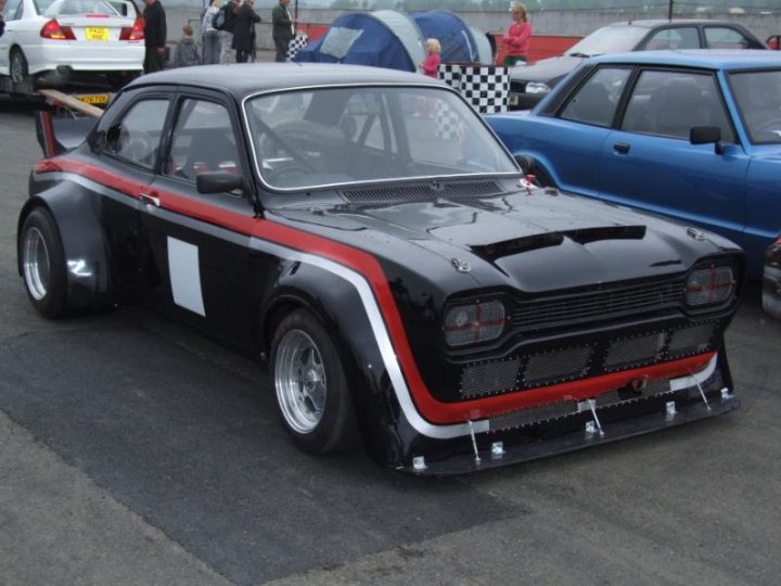 Modified Pistonheads Madness - The image shows a competition-style racing car on a track, characterized by its striking black and white livery with a red stripe. It has prominent features like a large rear wing, indicating it's designed for aerodynamics. In the background, there are other cars, indicating that this might be a race or a car show. The car is positioned on top of pavement, suggesting it's either parked or on display. The setting appears to be outdoors, and there are spectators around, suggesting it might be open to public viewing.