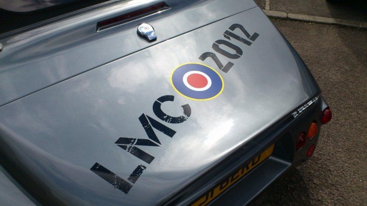 Le Mans classic stickers - Page 1 - South West - PistonHeads - The image depicts the back of a car with a distinctive year mark "2012" painted prominently, suggesting it's a military vehicle. Just below this, a circular logo with a red, white, and blue design includes the letters "LMGC," indicating the model or brand of the vehicle. The car appears to be parked in a lot, and the focus of the image is on the rear of the vehicle, capturing a close-up view of the text and logo. The photograph's composition and the objects within it give the impression of a calm, likely military-related event or vehicle showcase.