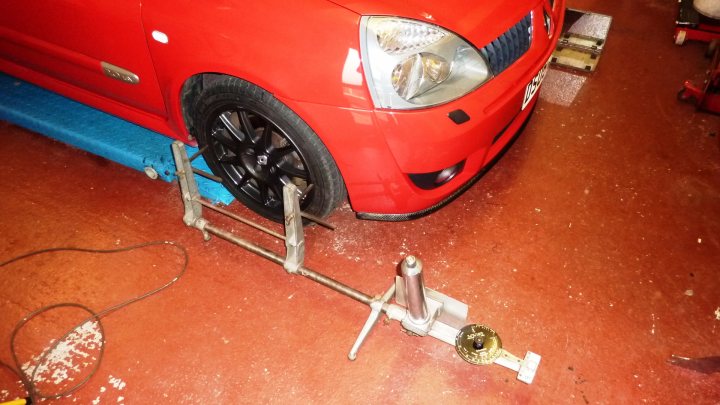 Suspension Alignment - Page 1 - Home Mechanics - PistonHeads - The image shows a close-up of the front wheel of a red car, which is likely in the process of an alignment or vehicle service. The car is stationed on a blue lift platform or serving rack, commonly used in shops or workshops to raise vehicles off the ground. Directly under the front wheel, there's a two-post car lift, suggesting the vehicle is being hoisted and the front wheel lowered onto the lift platform to expose the suspension or tires more easily. A silvertone stylus rests on a disc brake caliper, possibly a tool being used for diagnostics or maintenance. The background appears to be a work area with traditional automotive tools and equipment, indicating the setting might be a professional repair shop or garage.