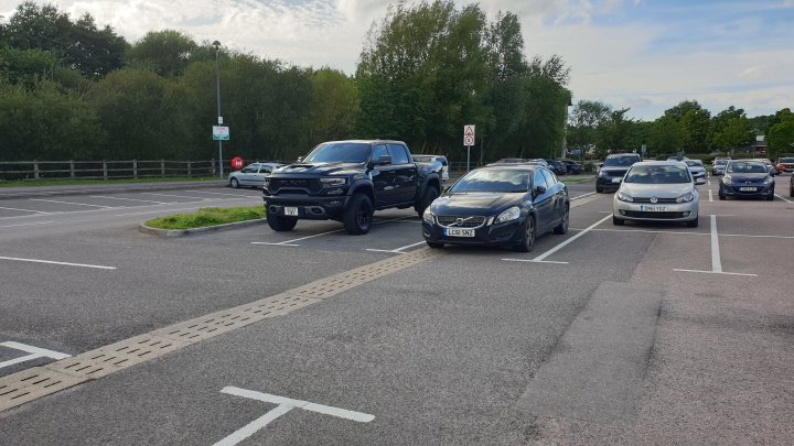 The BAD PARKING thread [vol4] - Page 648 - General Gassing - PistonHeads UK - The image shows an outdoor scene with a parking lot. There are multiple cars parked in the lot, and some vehicles appear to be in motion on the road adjacent to the parking lot. The sky above is overcast with clouds. In the background, there's vegetation that looks like it could be a grassy area or possibly a small forest. There is no visible text in the image. The style of the photo seems candid and may have been taken from inside a vehicle.