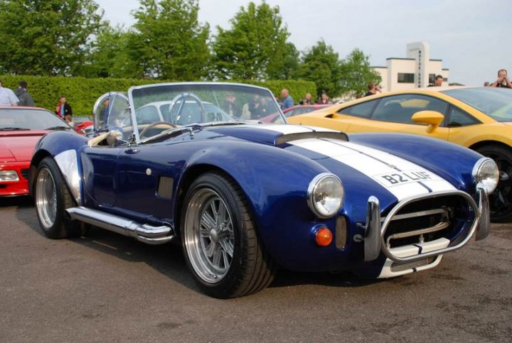 Sunday Supercar Pistonheads - The image showcases a vibrant scene of a rally car event in a parking lot. The focal point of the image is a sleek, blue, sporty CV22 Can-Am race car, parked on the asphalt road. This Can-Am has a prominent stripe down its center, adding to its striking appearance. Its round headlights and unique rear spoiler design suggest a focus on speed and aerodynamics. 

In the background, other cars are parked, including a vibrant red sports car. There are people gathered around the cars, adding a lively atmosphere to the scene. The lot is lush with green trees, providing a natural backdrop to the event. The image captures the excitement and allure of motorsports and car enthusiast gatherings.